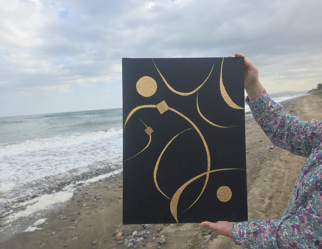 Person holding a black canvas with gold abstract designs on a beach.